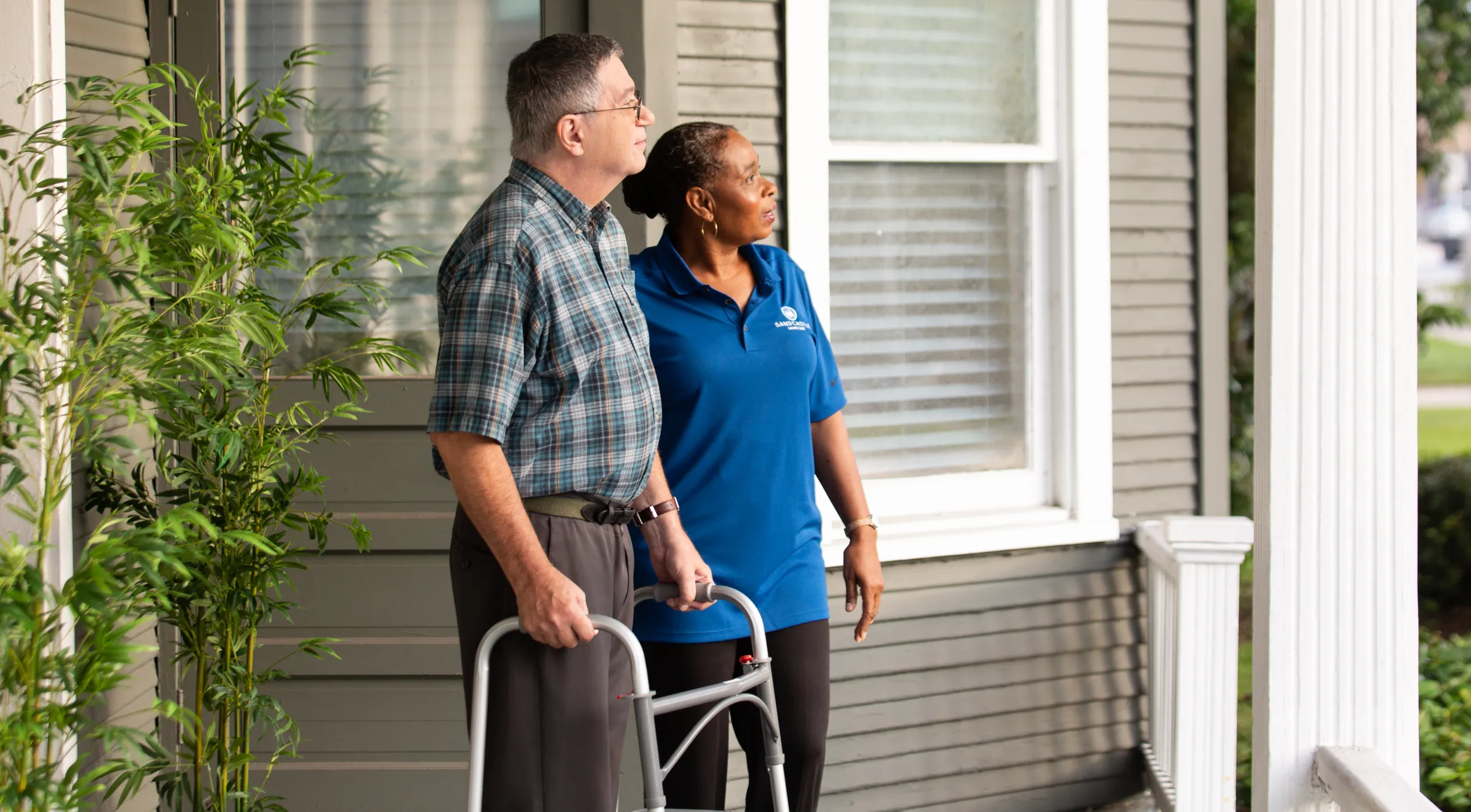 A caregiver walking beside an older man with a walker on his front porch