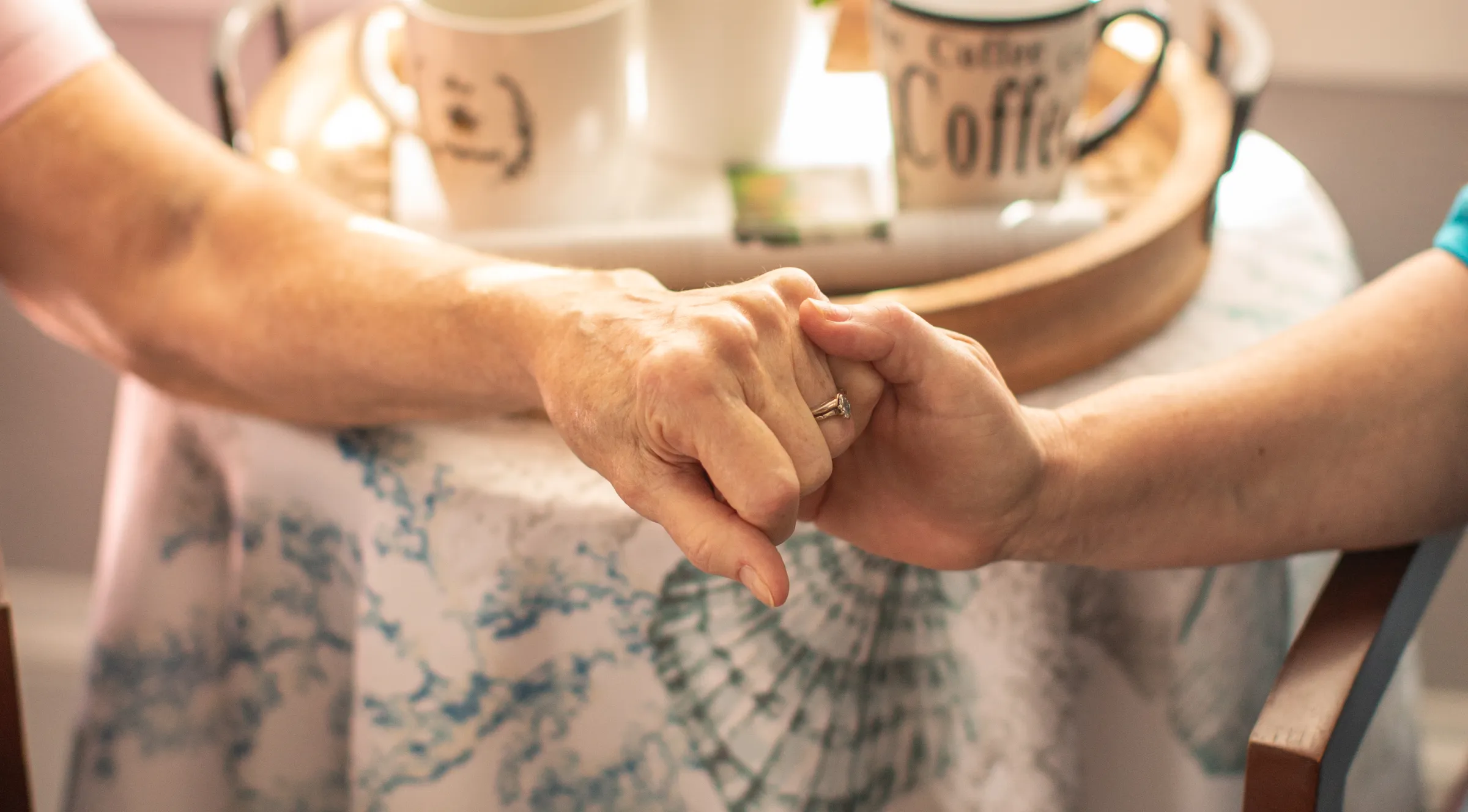 A caregiver holding an older woman's hands across a kitchen table
