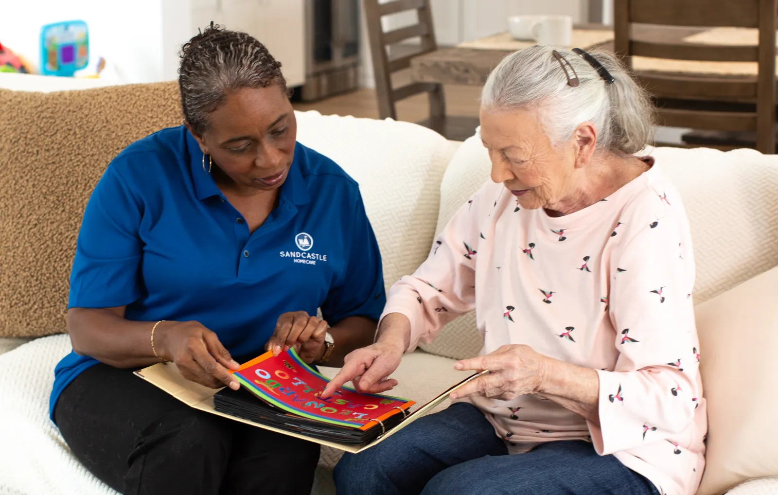 A caregiver and an older woman looking through a magazine together on the sofa